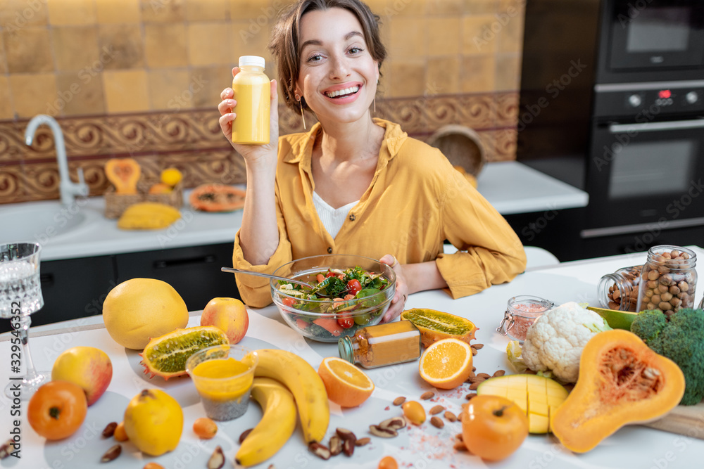 Portrait of a young and joyful woman holding bottle of juice at the table full of healthy raw vegeta