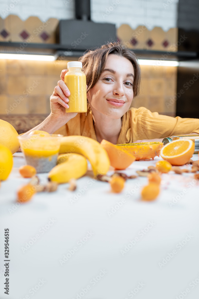 一个年轻快乐的女人的画像，她拿着一瓶果汁坐在装满健康生蔬菜的桌子旁