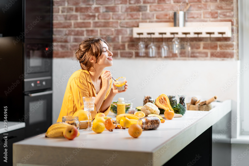 Cheerful woman eating chia pudding at the table full of healthy raw vegetables and fruits on the kit