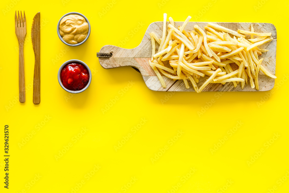 Fast food restaurant concept. French fries on cutting board near sauces on yellow table top-down cop