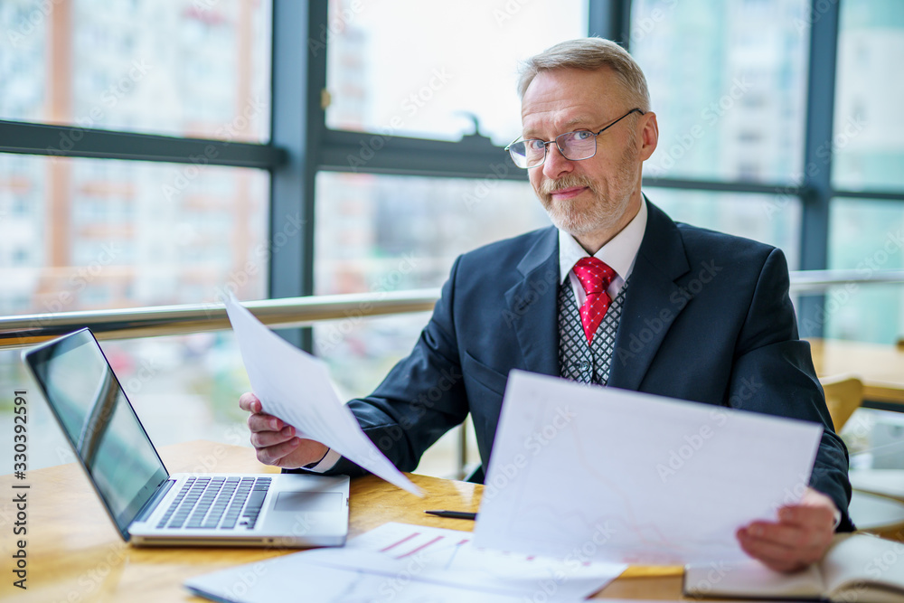 Mature businessman looking at laptop reading printed documents. Sitting near the window in office. P