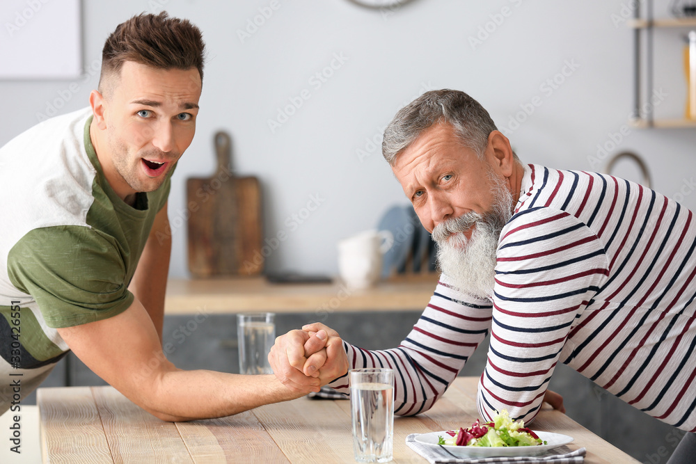 Senior man and his adult son having arm wrestling competition in kitchen