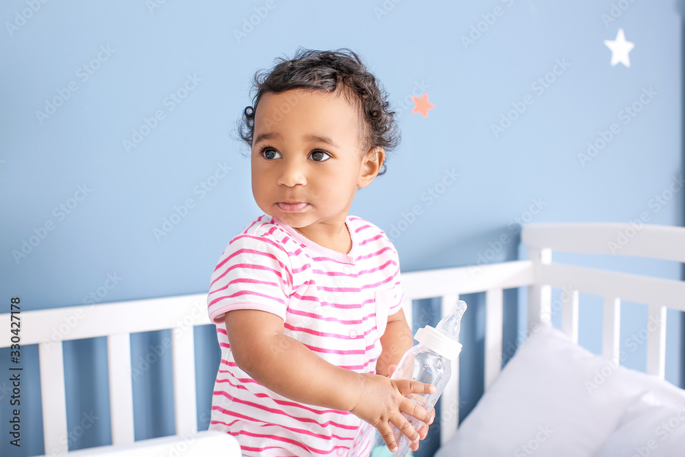 Cute African-American baby with bottle of water in crib