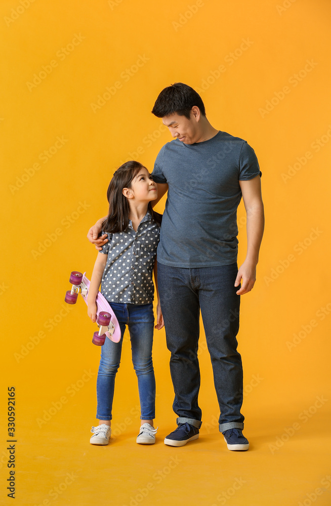 Happy Asian man and his little daughter with skateboard on color background