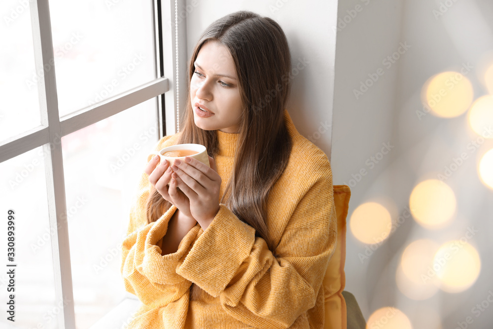 Beautiful young woman with cup of tea sitting on window sill at home