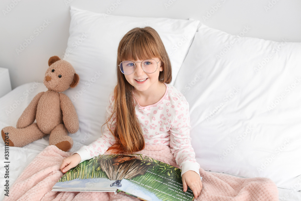 Cute little girl wearing glasses while reading book in bed