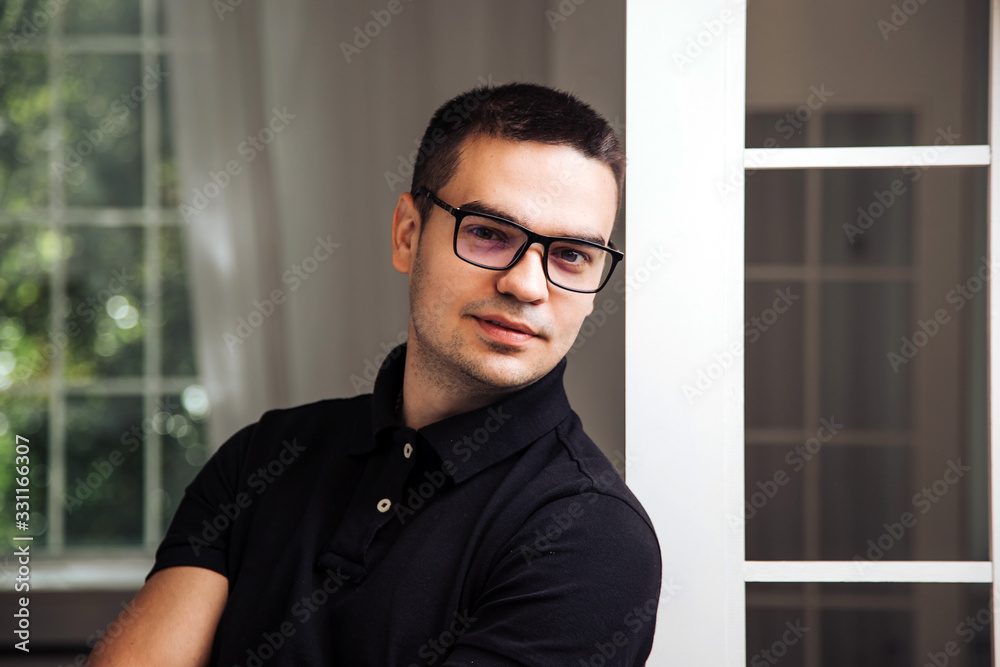 Adult man in a black shirt in eyeglasses with black rim. Male poses near window. Close-up