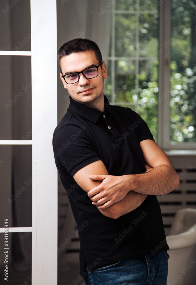 Handsome man in trendy glasses and black shirt looks away on the background of window in the room. P