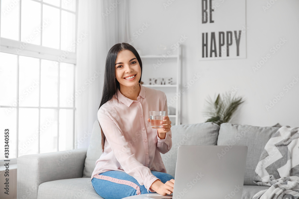 Beautiful young woman drinking water while using laptop at home