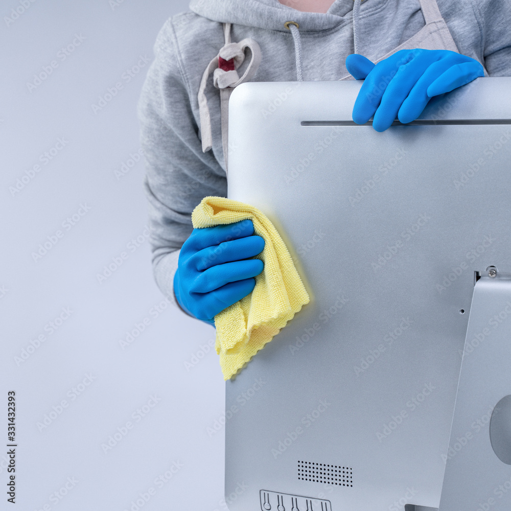 Young woman housekeeper in apron is cleaning silver computer surface with blue gloves, wet yellow ra