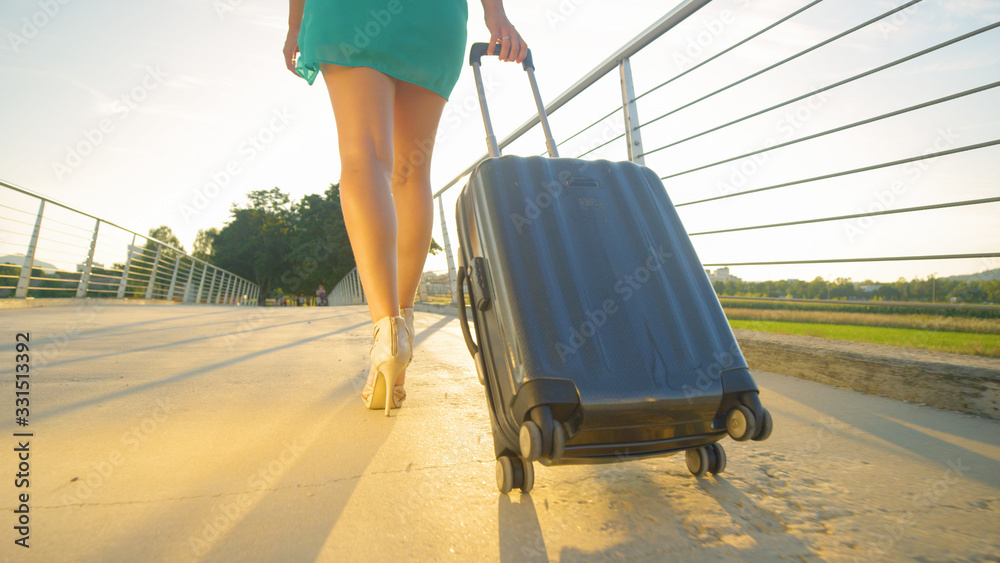 LOW ANGLE: Pretty woman carries her travel luggage across a bridge at sunrise.