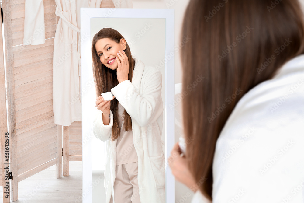 Beautiful young woman applying cream near mirror