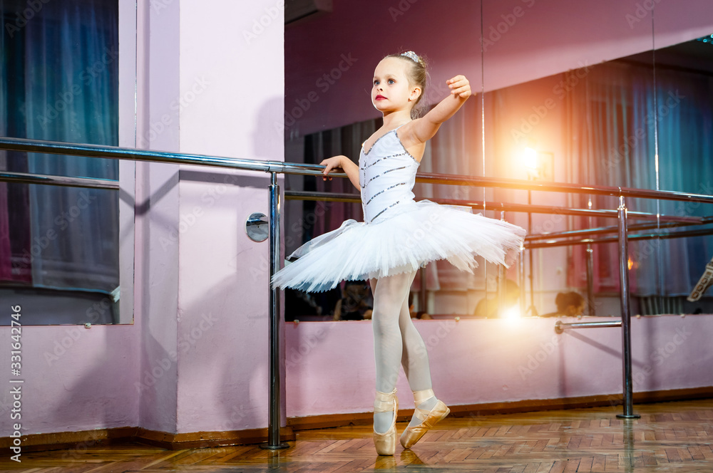 Small ballerina girl in white tutu standing next to the mirror with her reflection in it at dance st