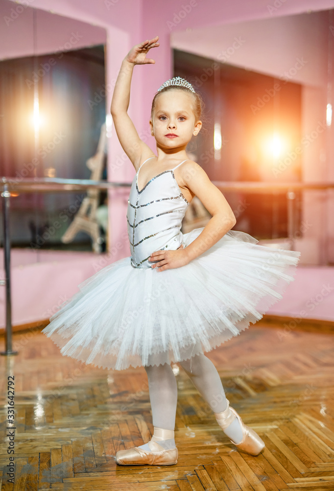 Little adorable ballerina girl in a white tutu standing in pose near the mirror and looking at the c