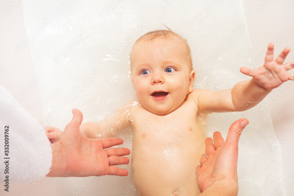 Laughing baby splash on the back in bathroom tub at home reaching for mother hands