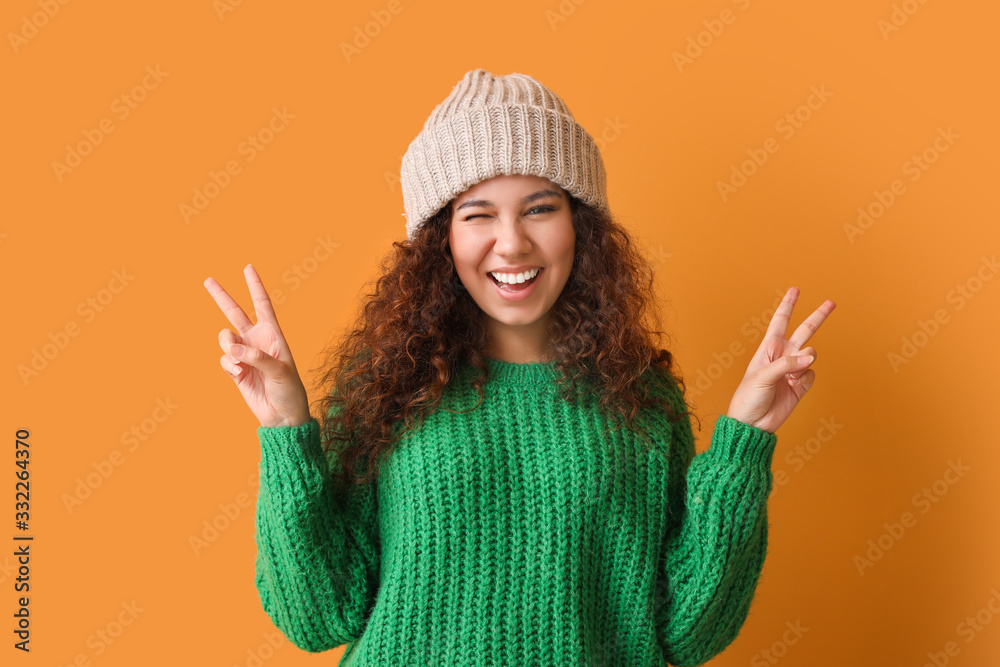 Young woman in warm sweater showing victory gesture on color background