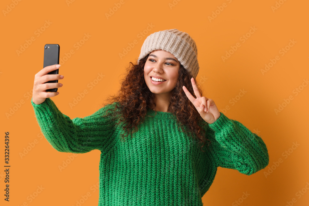 Young woman in warm sweater taking selfie on color background