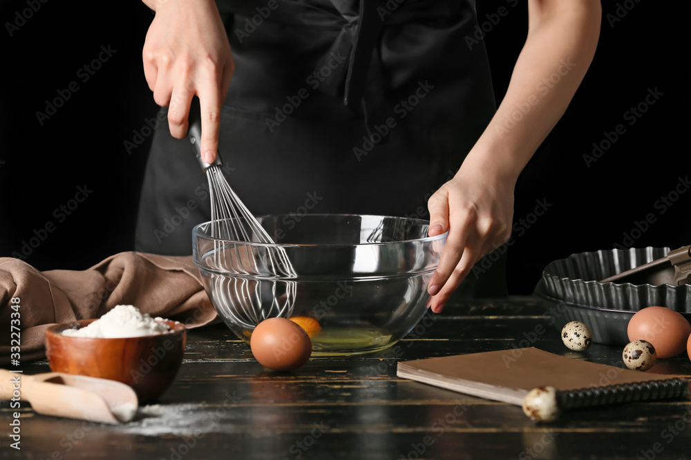 Woman with cook book making dough at table on dark background