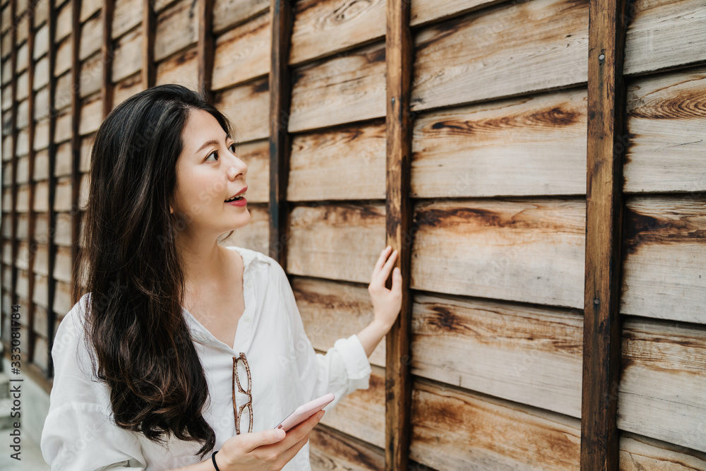 curious young asian japanese lady tourist walking along wooden wall and holding cellphone with onlin