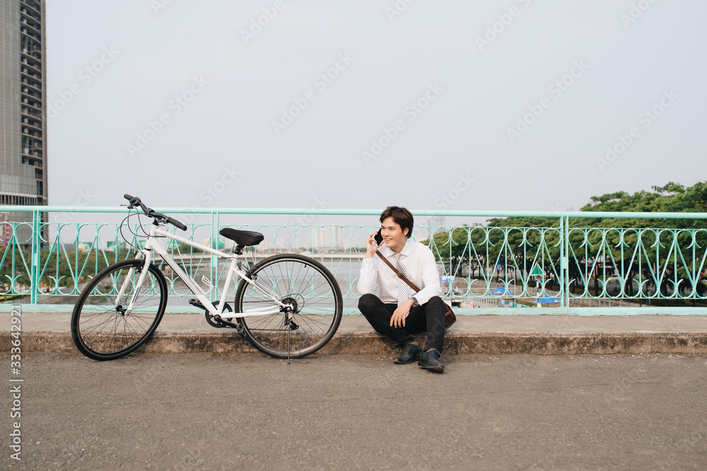 Young asian man talking by mobile phone while sitting on the ground near the bike, outdoors. Wearing