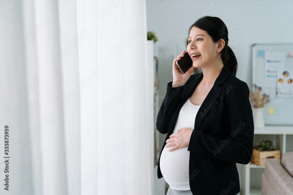 Pregnant office lady in formal suit communicating on cellphone by window. Cheerful motherhood busine