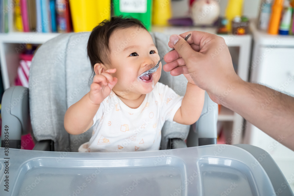 Father feed mashed food to Asian baby cute girl who sit in highchair at home. Little girl look at da