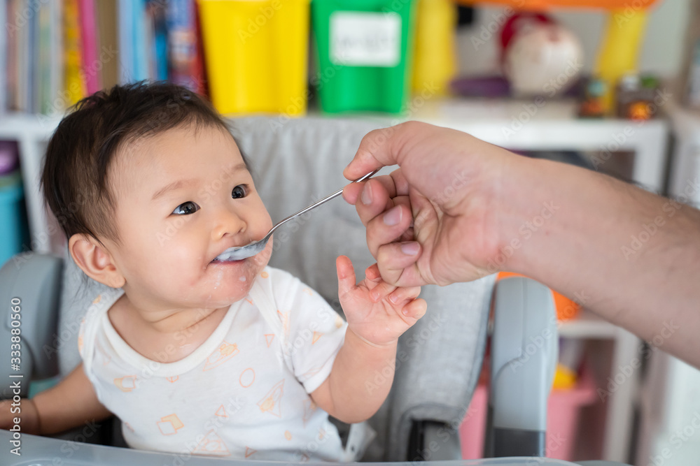 Father feed mashed food to Asian baby cute girl who sit in highchair at home. Little girl look at da