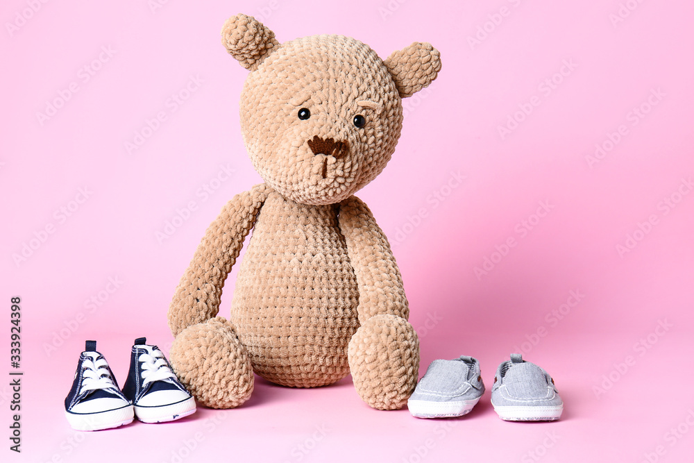 Teddy bear with baby shoes on color background