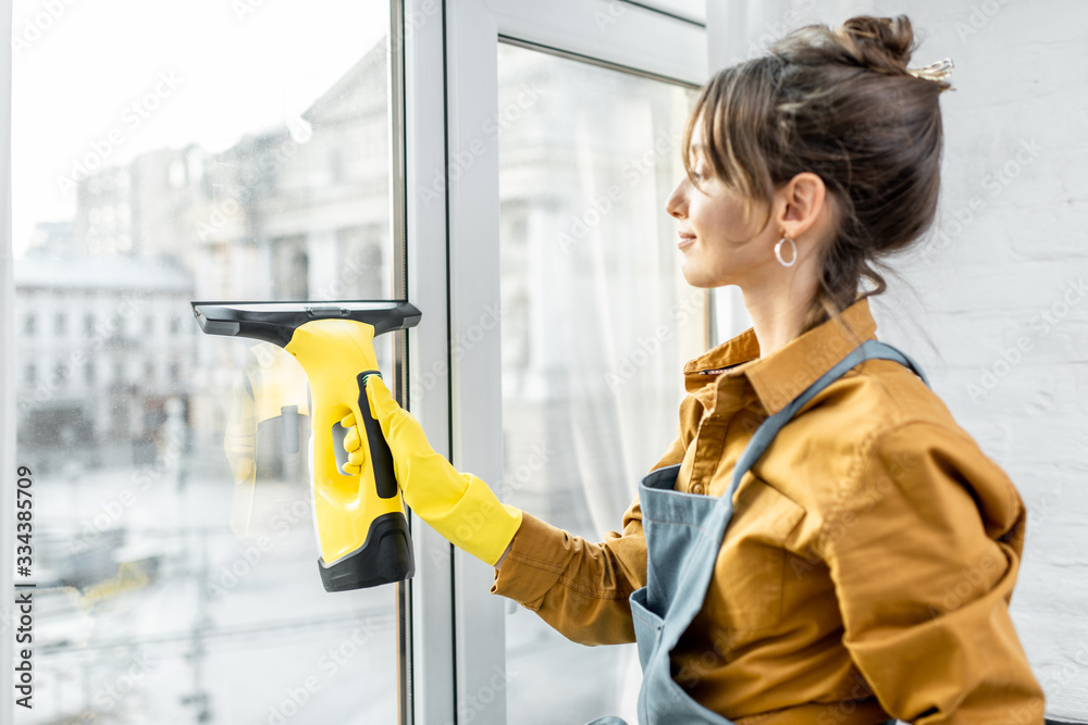 Beautiful young housewife in working clothes washing windows by professional tools at home