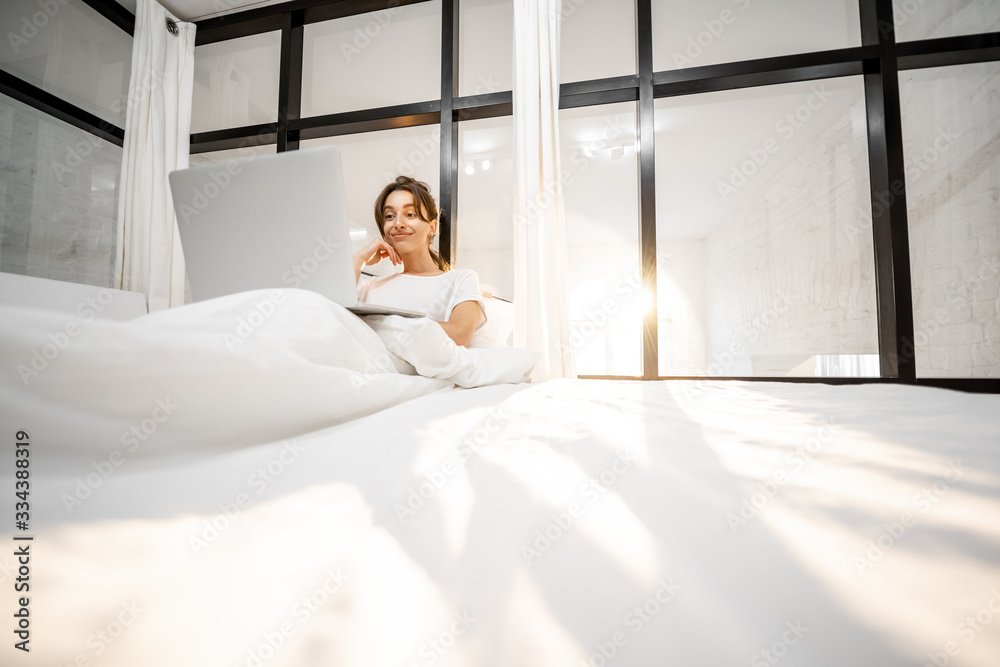 Woman working on a laptop, lying in the beautiful white and sunny bedroom, wide interior view with c