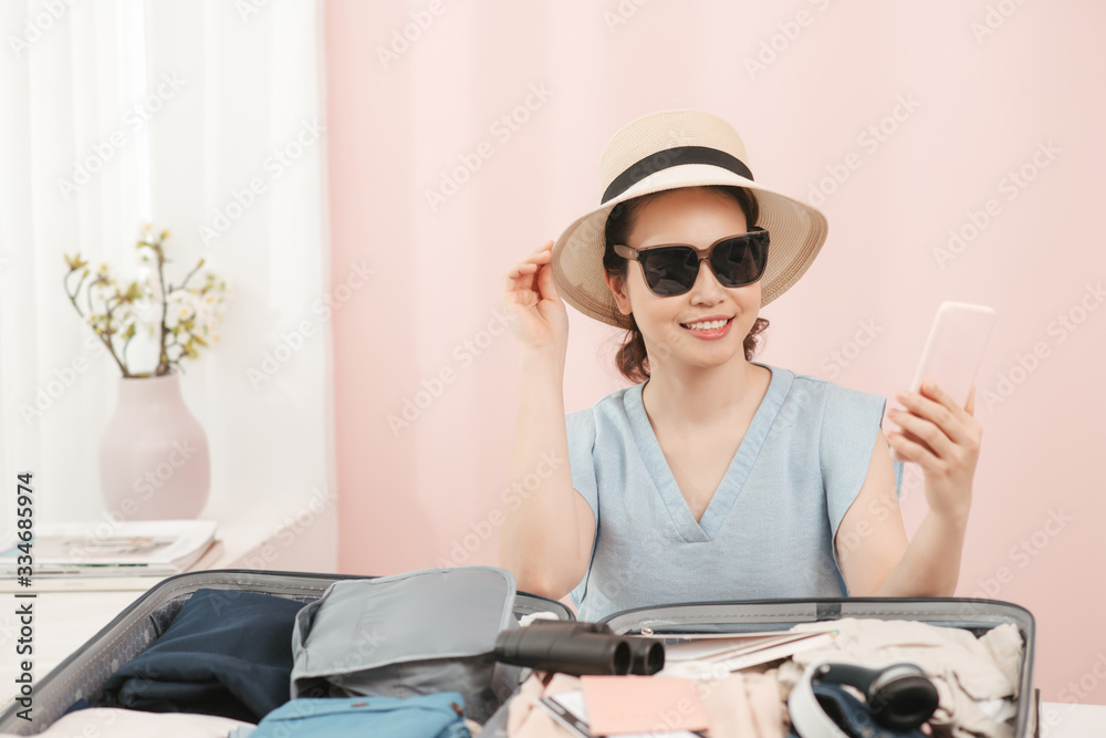 Young woman choosing hat sitting with clothes and suitcase on the bed. Prepairing for the summer vac