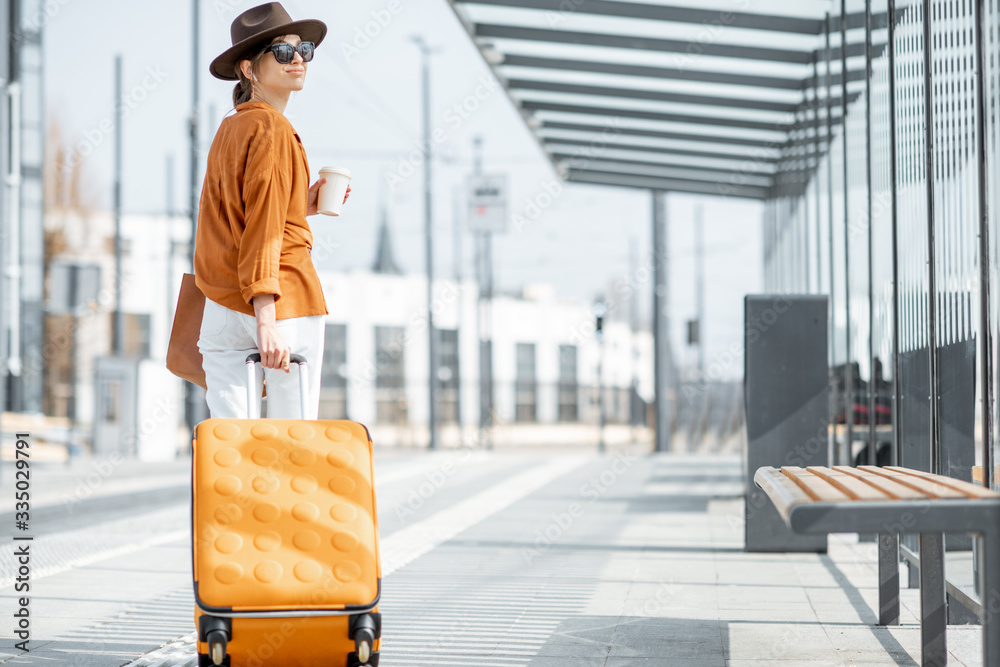 年轻的女性旅行者提着黄色行李箱在户外现代交通站行走，背后的生活