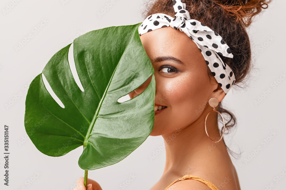 Beautiful African-American woman with tropical leaf on light background