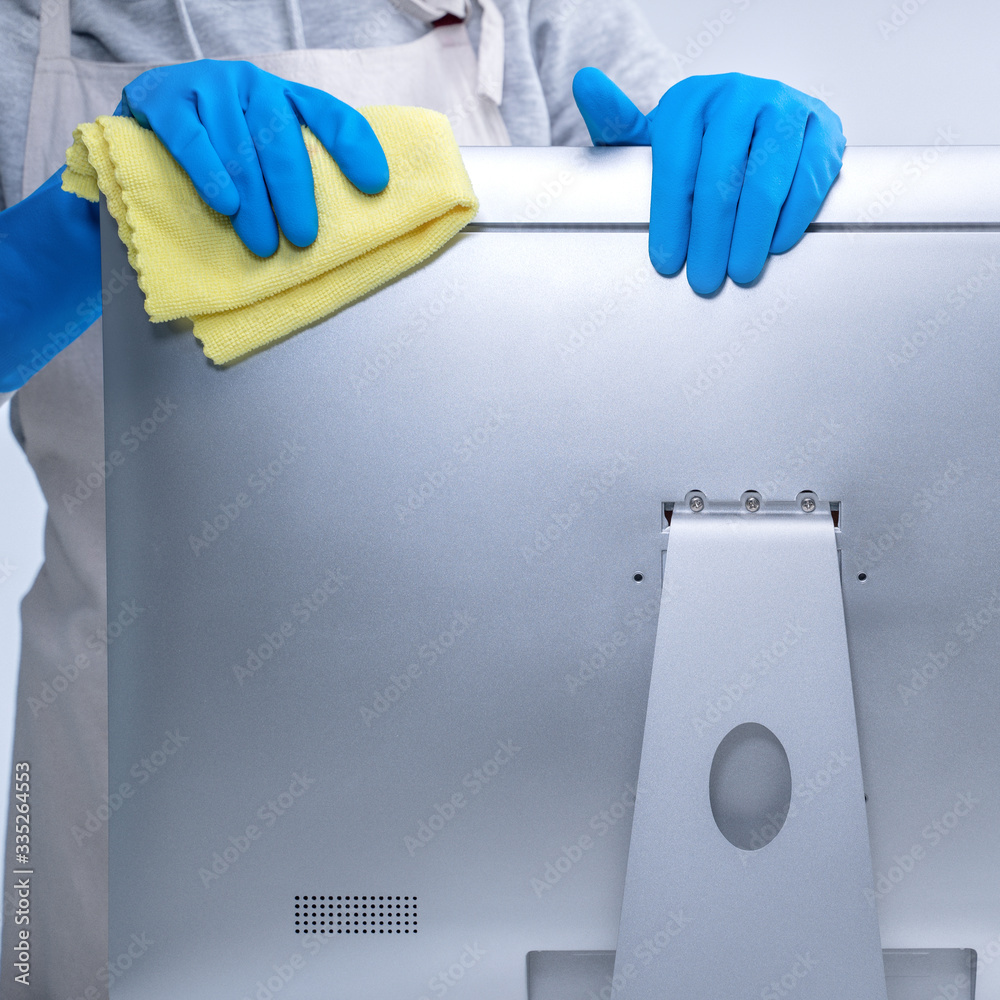 Young woman housekeeper in apron is cleaning silver computer surface with blue gloves, wet yellow ra