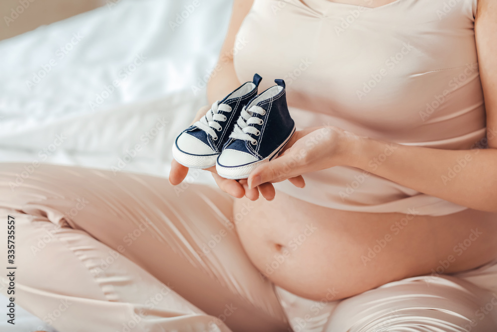 Beautiful pregnant woman with baby shoes in bedroom, closeup