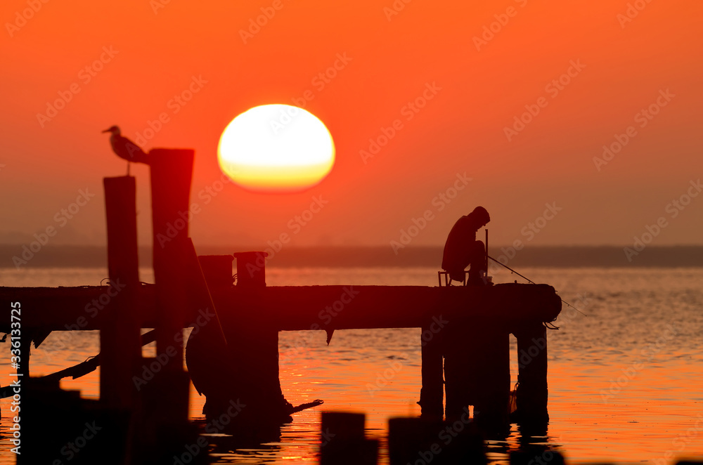 Silhouette of a fisherman and a bird  in the rays of the rising sun and gentle red light