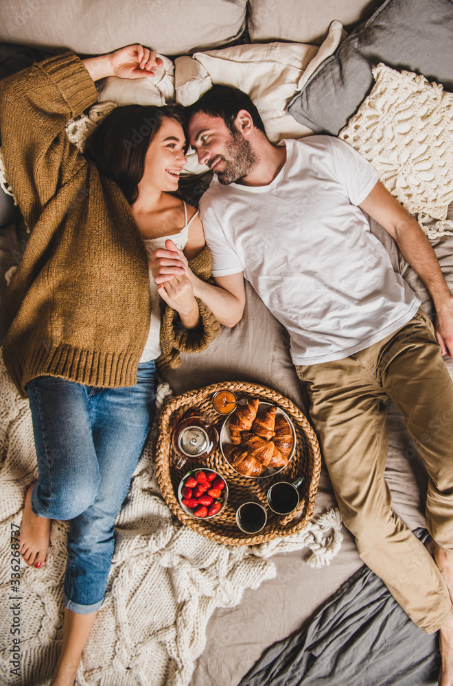 Young smiling happy couple in love lying on pastel colored blankets barefoot in bed, having breakfas