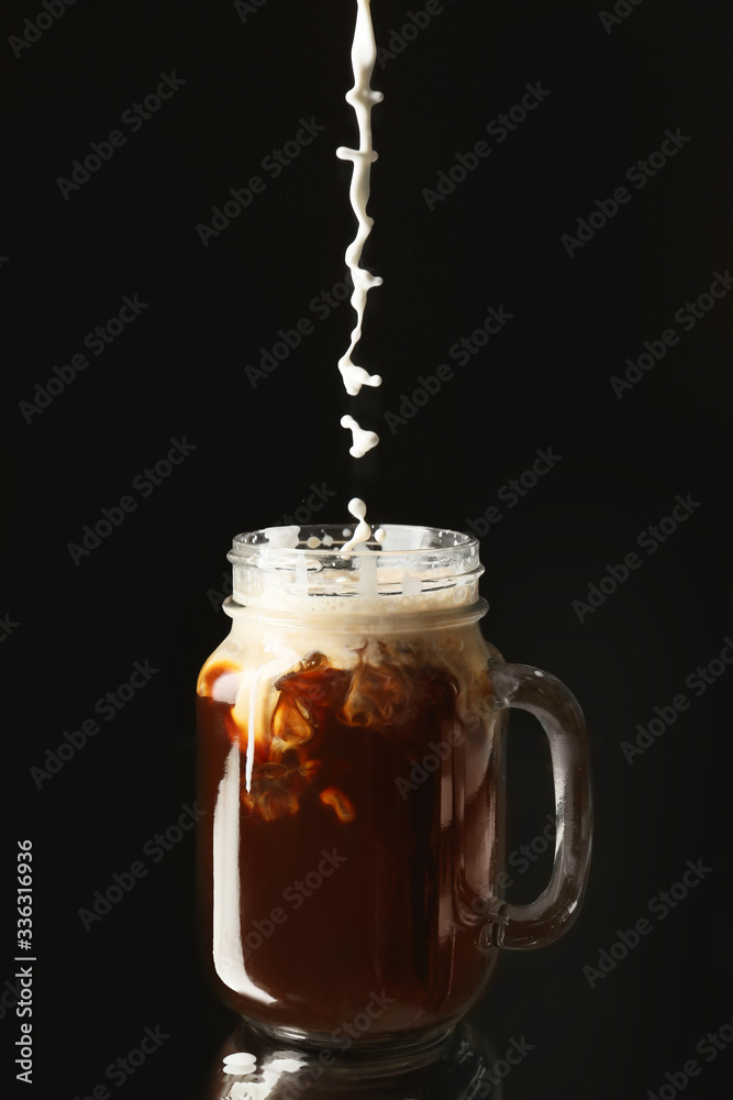 Pouring of milk into cold coffee in mason jar on dark background