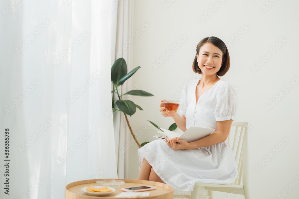 Young woman at home sitting near window relaxing in her living room reading book and drinking tea