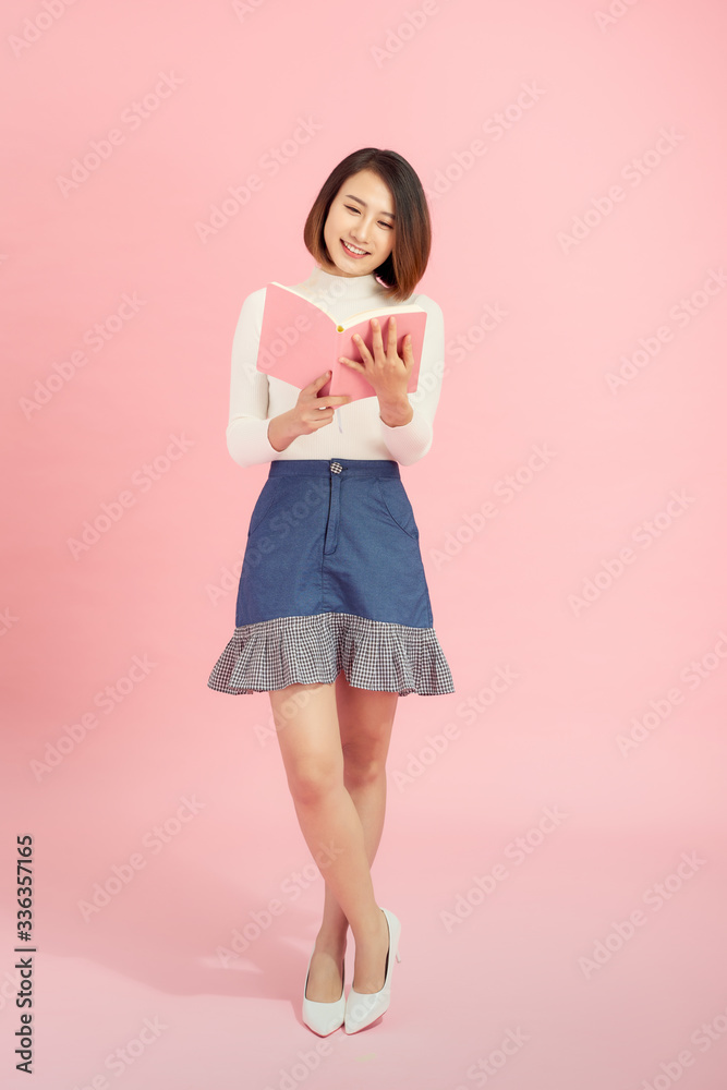 Full length portrait of a smiling beautiful Asian woman student holding book isolated on a pink back