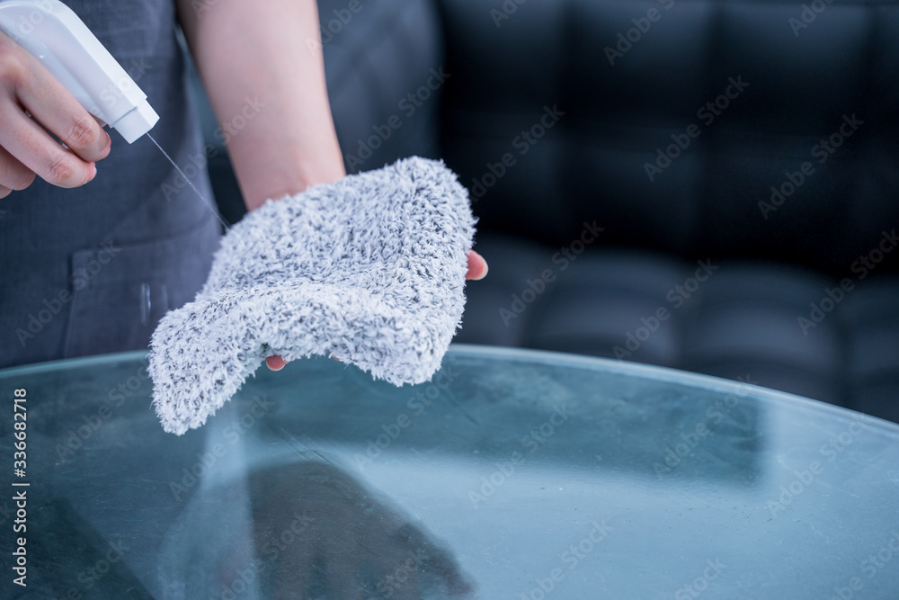 Young woman maid in apron is cleaning, wiping down office glass table surface with spray bottle clea