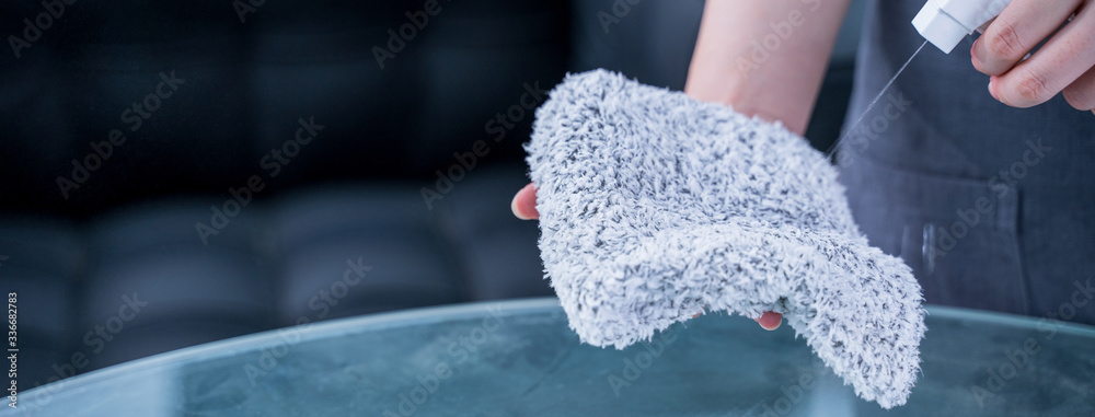 Young woman maid in apron is cleaning, wiping down office glass table surface with spray bottle clea