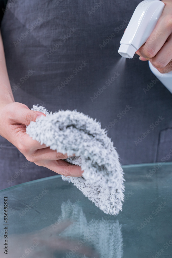 Young woman maid in apron is cleaning, wiping down office glass table surface with spray bottle clea