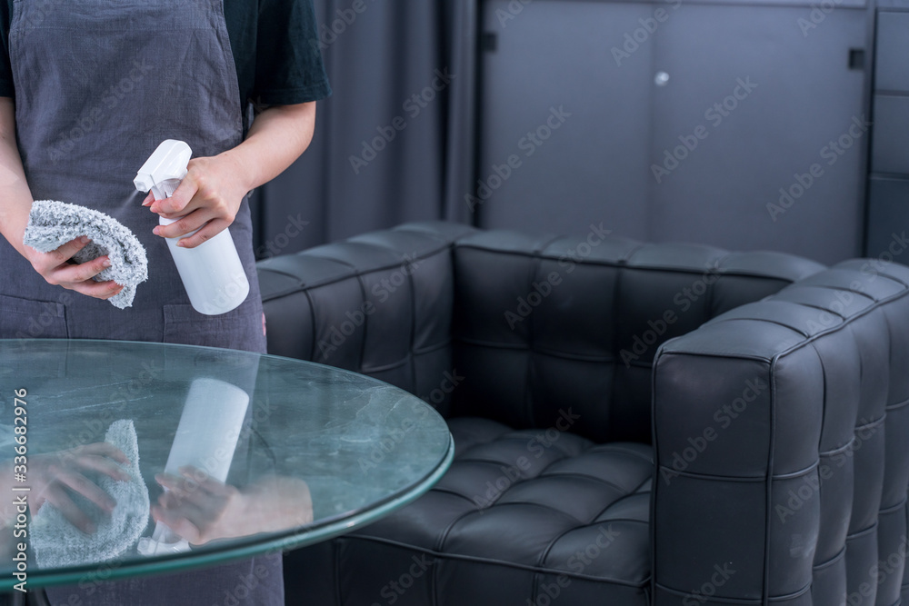 Young woman maid in apron is cleaning, wiping down office glass table surface with spray bottle clea