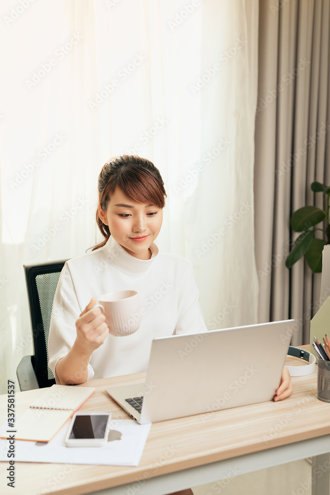 漂亮的年轻亚洲女人在家用笔记本电脑工作时喝茶或喝咖啡