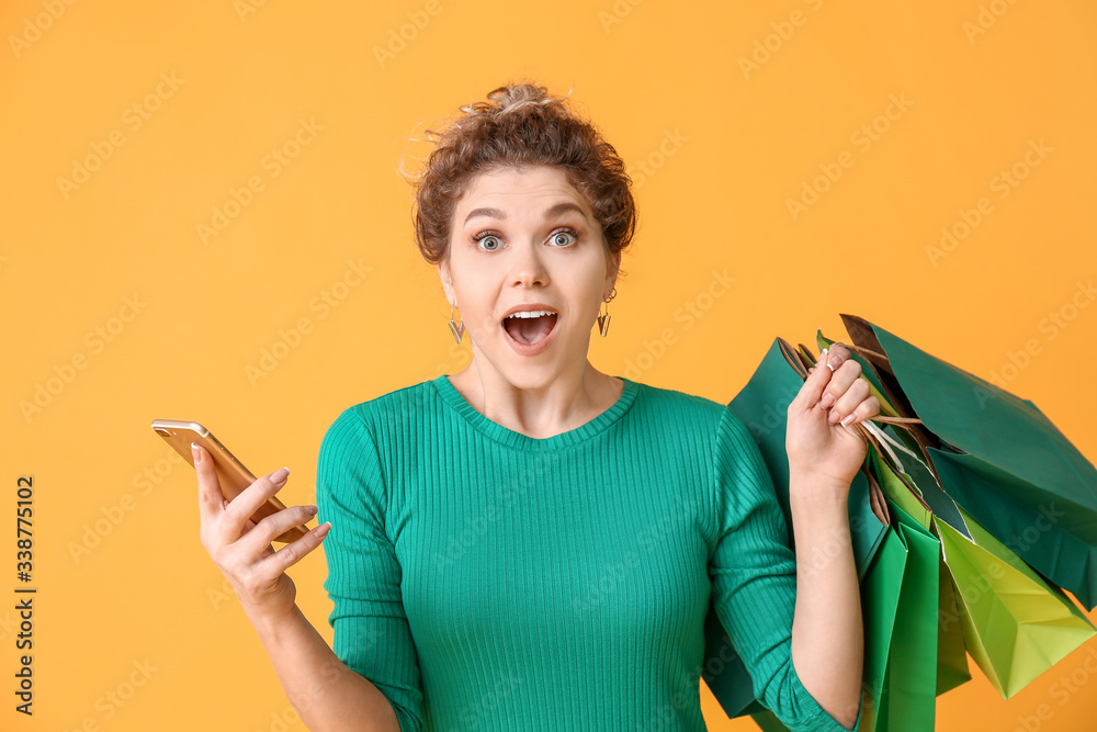 Surprised young woman with mobile phone and shopping bags on color background