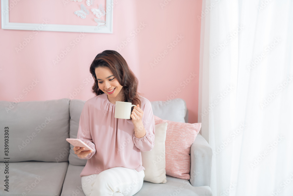 Attractive young Asian woman using phone and drinking coffee when sitting on sofa at home.