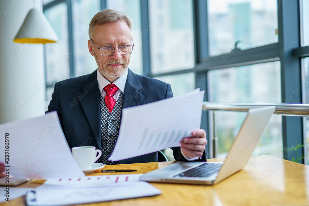 Thoughtful middle aged businessman in suit with a laptop sitting near the window working with docume