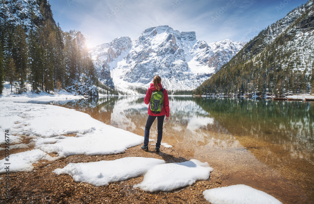 Young woman with backpack on the snowy shore of Braies lake with clear water at sunny bright day in 