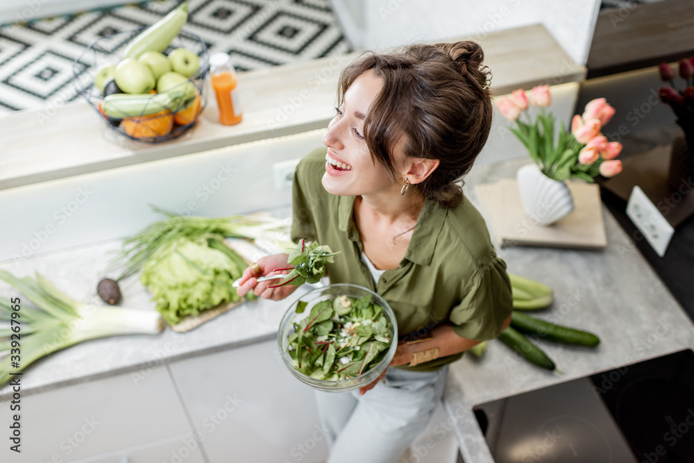 Portrait of a young and cheerful woman eating salad standing on the kitchen with food ingredients on